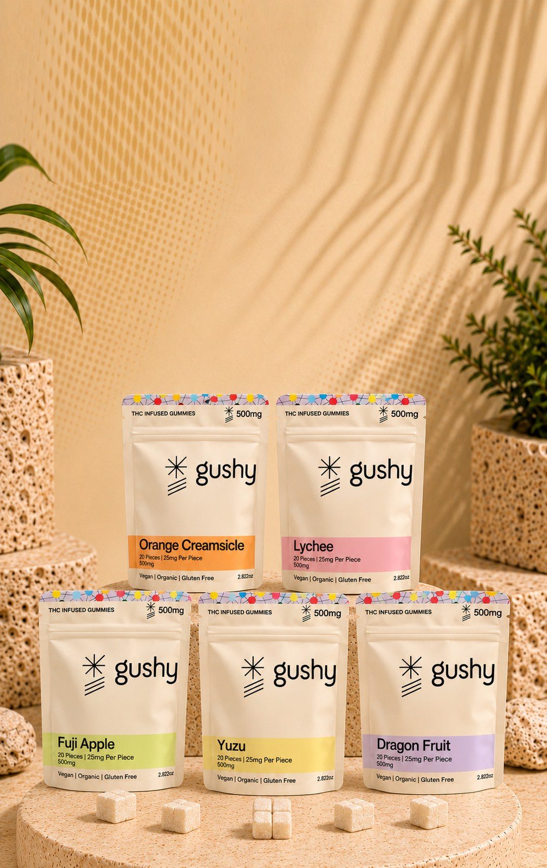 The five Gushy live resin gummy pouches, Fuji Apple, Orange Creamsicle, Yuzu, Lychee and Dragon Fruit, on a travertine platform with scored, sugar-dusted gummies in front